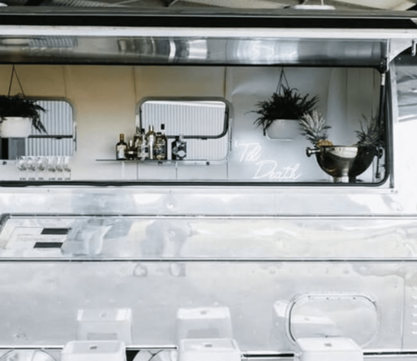 The polished exterior of the Airstream with the bar service window open, hanging ferns and the Til Death neon visible inside