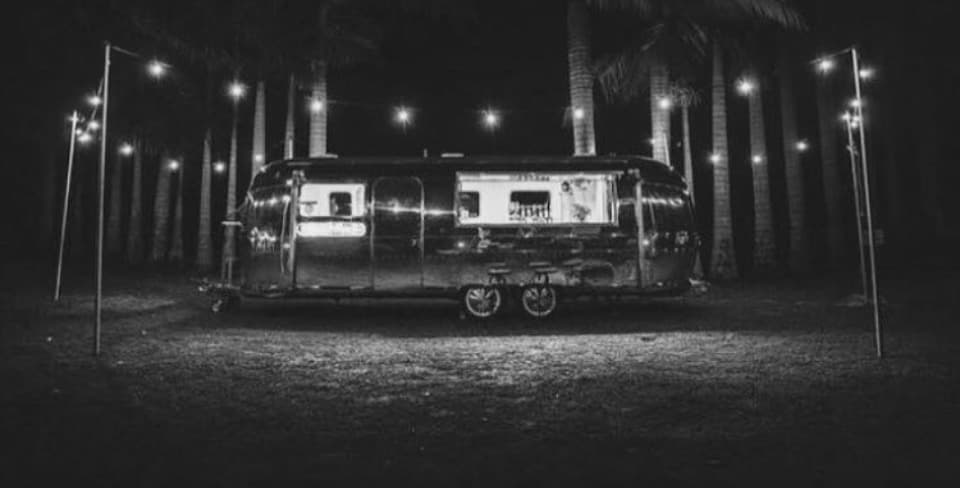 The Airstream glowing at night, framed by palm trees and festoon lighting