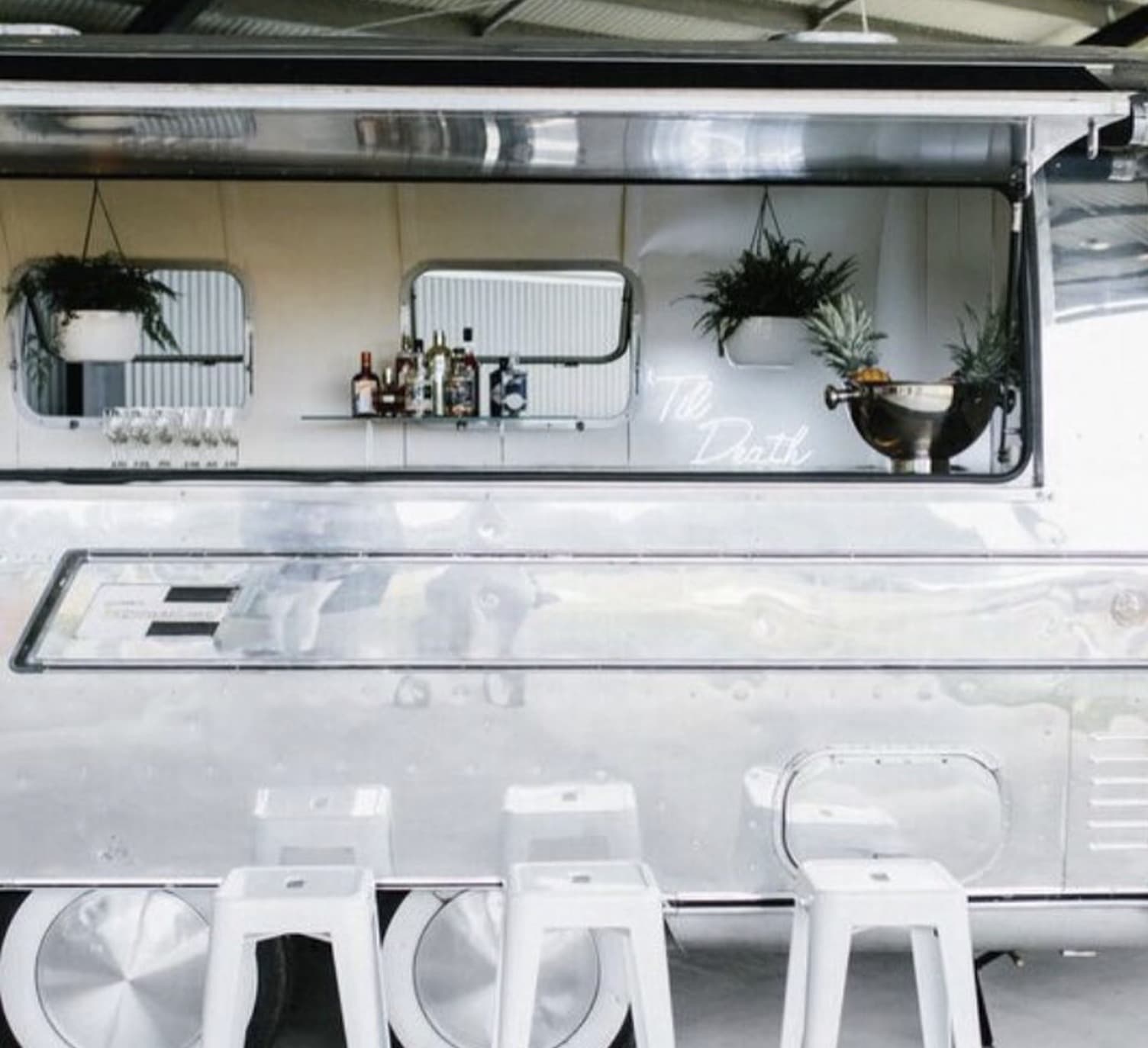The polished Airstream bar window open with three white bistro stools, hanging ferns and the Til Death neon visible inside