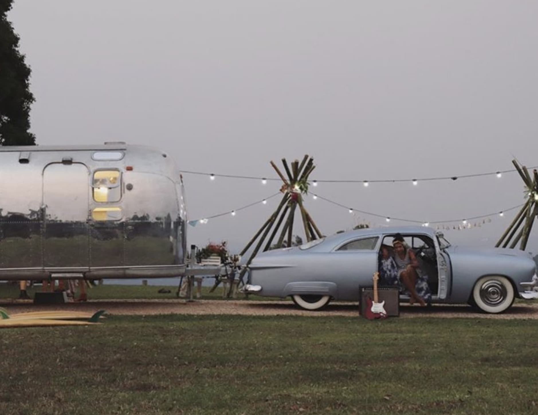 The polished Airstream alongside a vintage blue Cadillac at dusk, bamboo tipis and festoon lighting between them