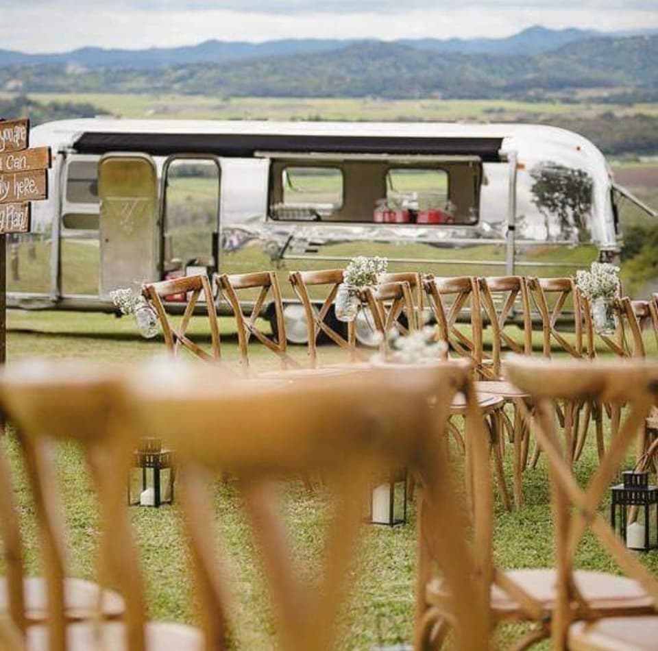 Ceremony aisle with crossback chairs and hanging florals, Airstream behind and hinterland view
