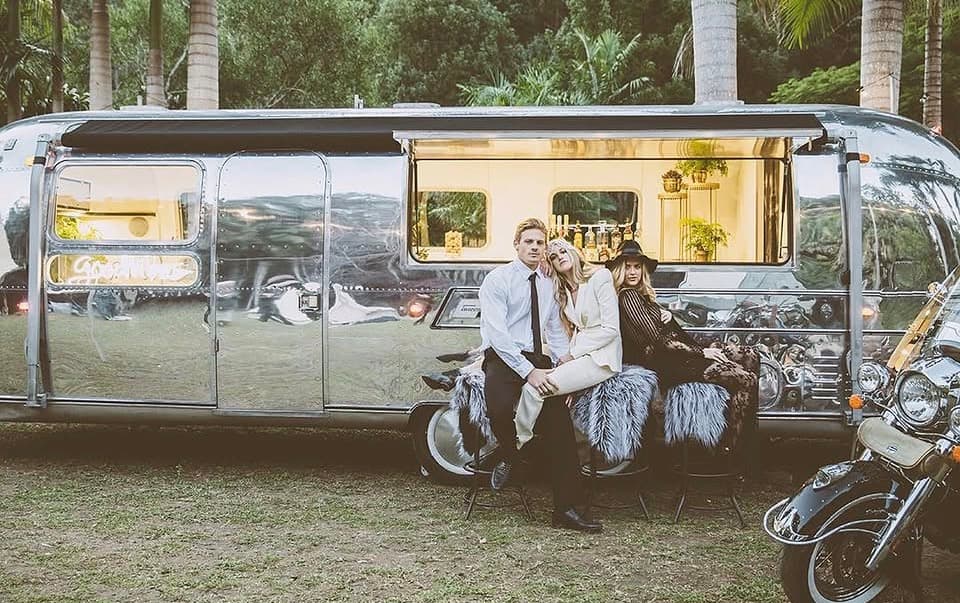 Couple seated on white fur stools in front of the polished Airstream, glowing interior and motorbike in frame